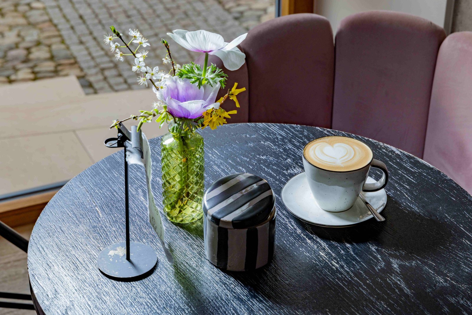 Cappuccino mit Herz-Latte-Art, frischen Blumen und grünem Glasväschen auf einem Café-Tisch