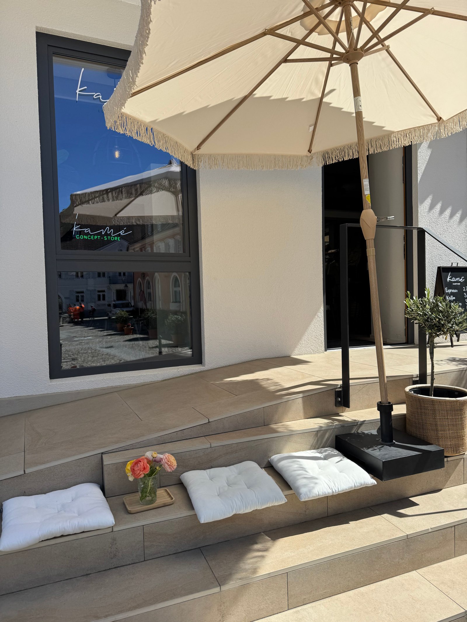 Kamé Store-Eingang mit Sonnenschirm, Sitzkissen und Blumen